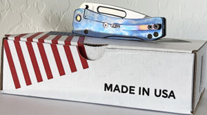 Blue anodized Medford Slim Midi knife on a red and white striped box. Visible blade and handle. Challenge Coin USA branding.