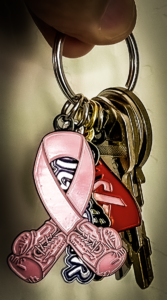 Pink breast cancer ribbon keychain hangs from silver key rings. Pink ribbon design with red lettering on a black background.